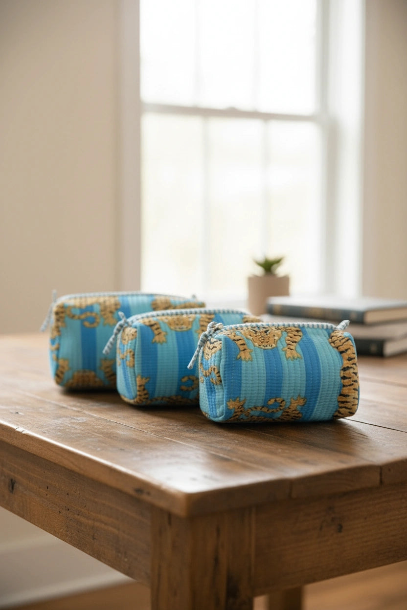 Three zippered cosmetic pouches with a blue and gold floral pattern, arranged on a rustic wooden table in a softly lit room.