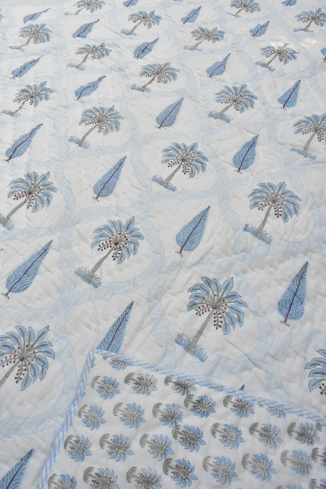 A close-up of a light blue and white quilted jaipuri design featuring embroidered palm trees and leaf motifs, with a braided border visible on the right.