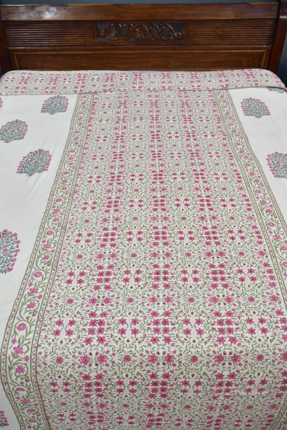 A floral patterned dohar / AC blanket with pink and green motifs on a cream background, laid out on a bed with a dark wooden headboard.