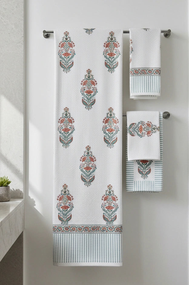 A set of white waffle-weave towels with a teal and rust orange botanical damask pattern, hanging on a silver metal towel bar against a white wall.