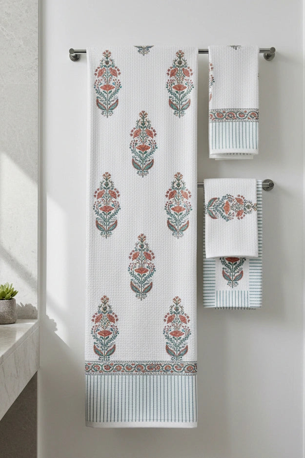 A set of white waffle-weave towels with a teal and rust orange botanical damask pattern, hanging on a silver metal towel bar against a white wall.