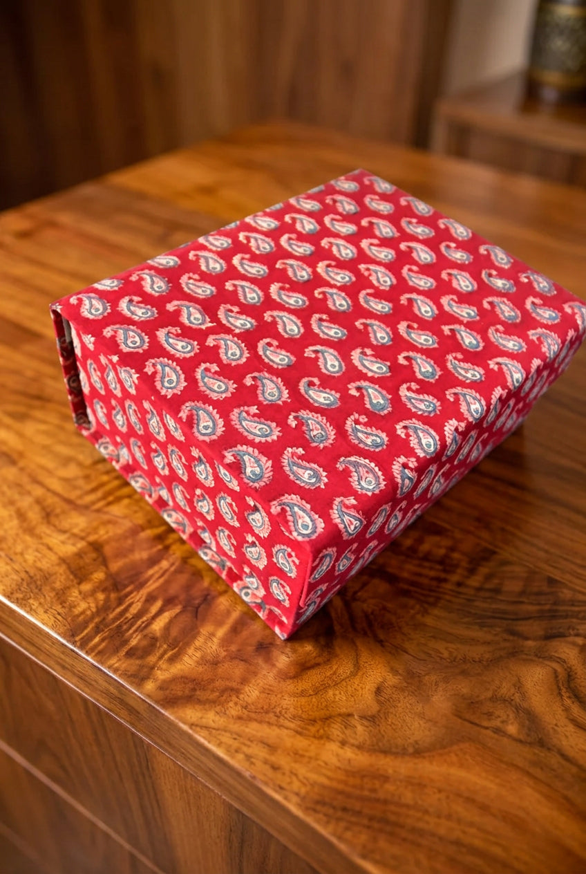 A rectangular gift box wrapped in red fabric with a white and blue paisley pattern, resting on a dark wooden surface.