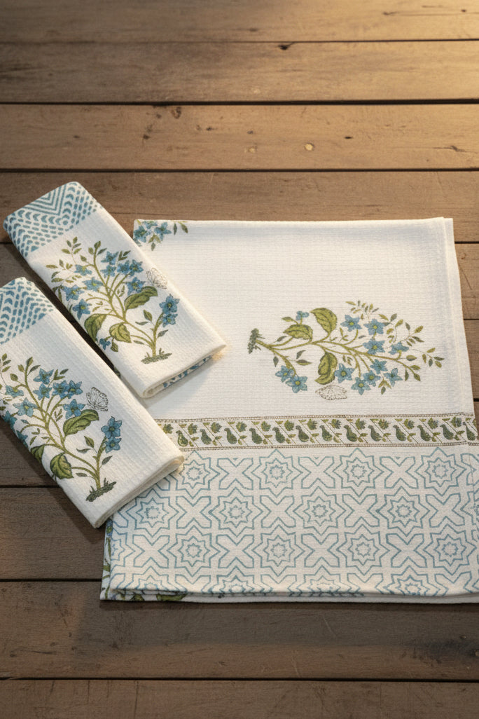 A set of three white cotton kitchen towels with blue and green floral embroidery and geometric patterns, laid out on a textured blue background.
