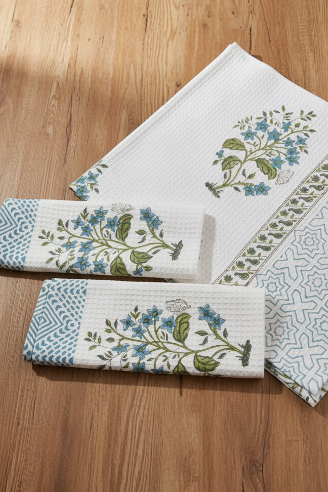 A set of white cotton napkins with blue floral prints, green leaves, and geometric borders, arranged on a textured blue background.