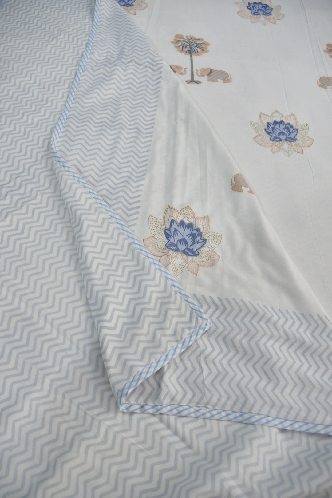 A close-up of a white dohar with a light blue and white chevron patterned border and matching blue-and-white stitched edge. The main body is white with scattered floral motifs in blue, beige, and muted green.