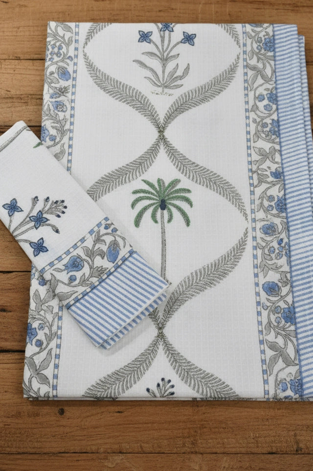 A folded white cotton fabric set, towel & napkin set, featuring blue floral prints, gray palm tree motifs, and blue-and-white striped borders, displayed on a rustic wooden surface.