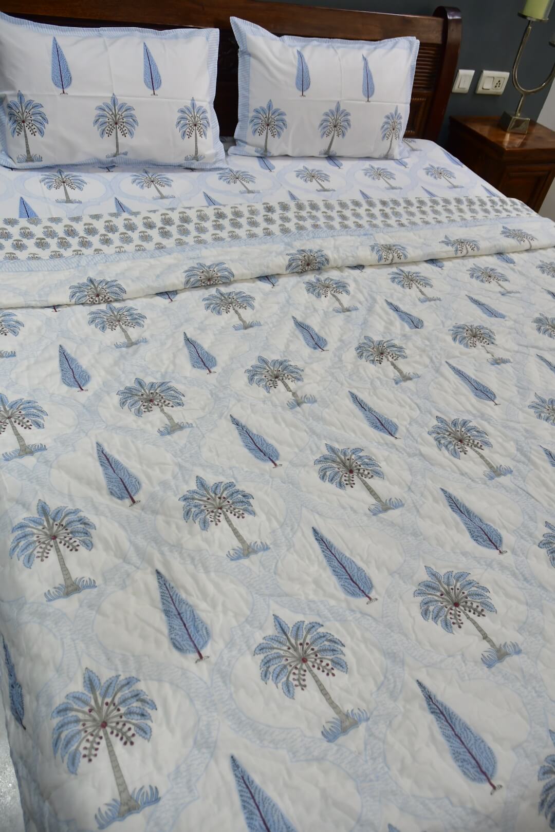 A bed dressed with a light blue and white quilted bedding set featuring a palm tree and feather pattern, with two matching pillowcases and a patterned runner, shown in a bedroom setting.