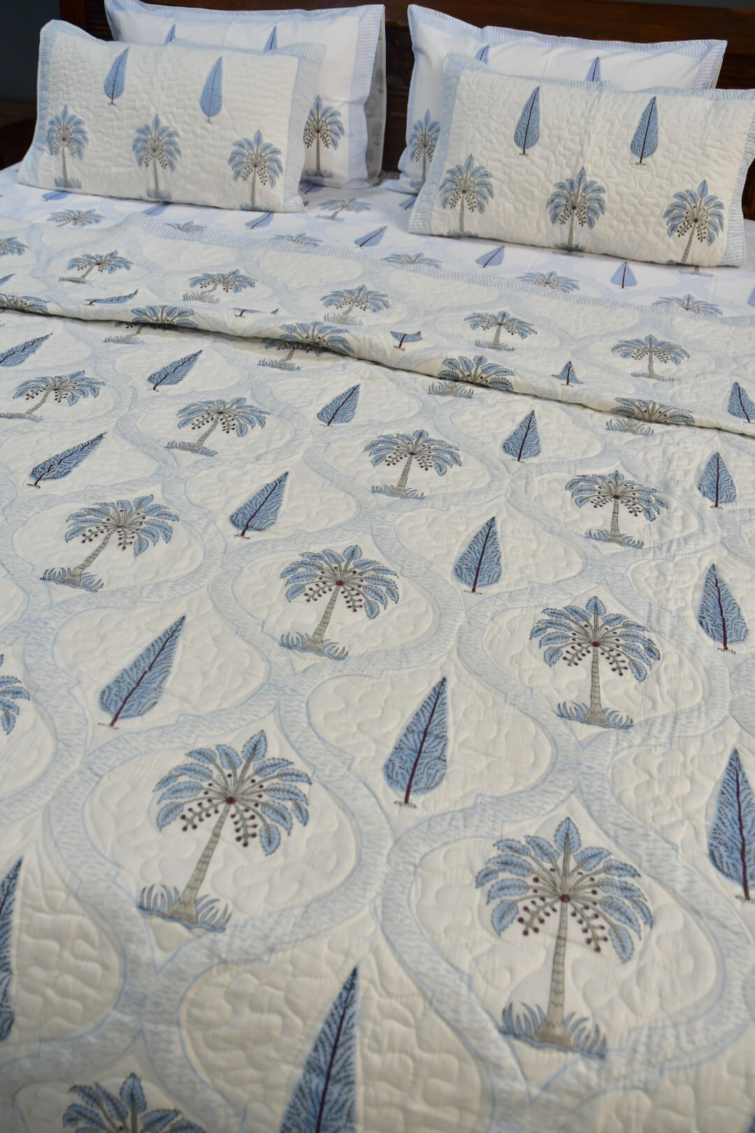 A quilted bedspread and matching pillow shams featuring a repeating pattern of palm trees and blue teardrop motifs on a white background, styled on a bed.