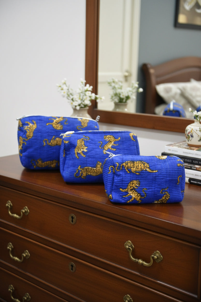 Three blue zippered pouches with a gold tiger pattern, arranged diagonally on a blue textured surface in a bedroom setting.