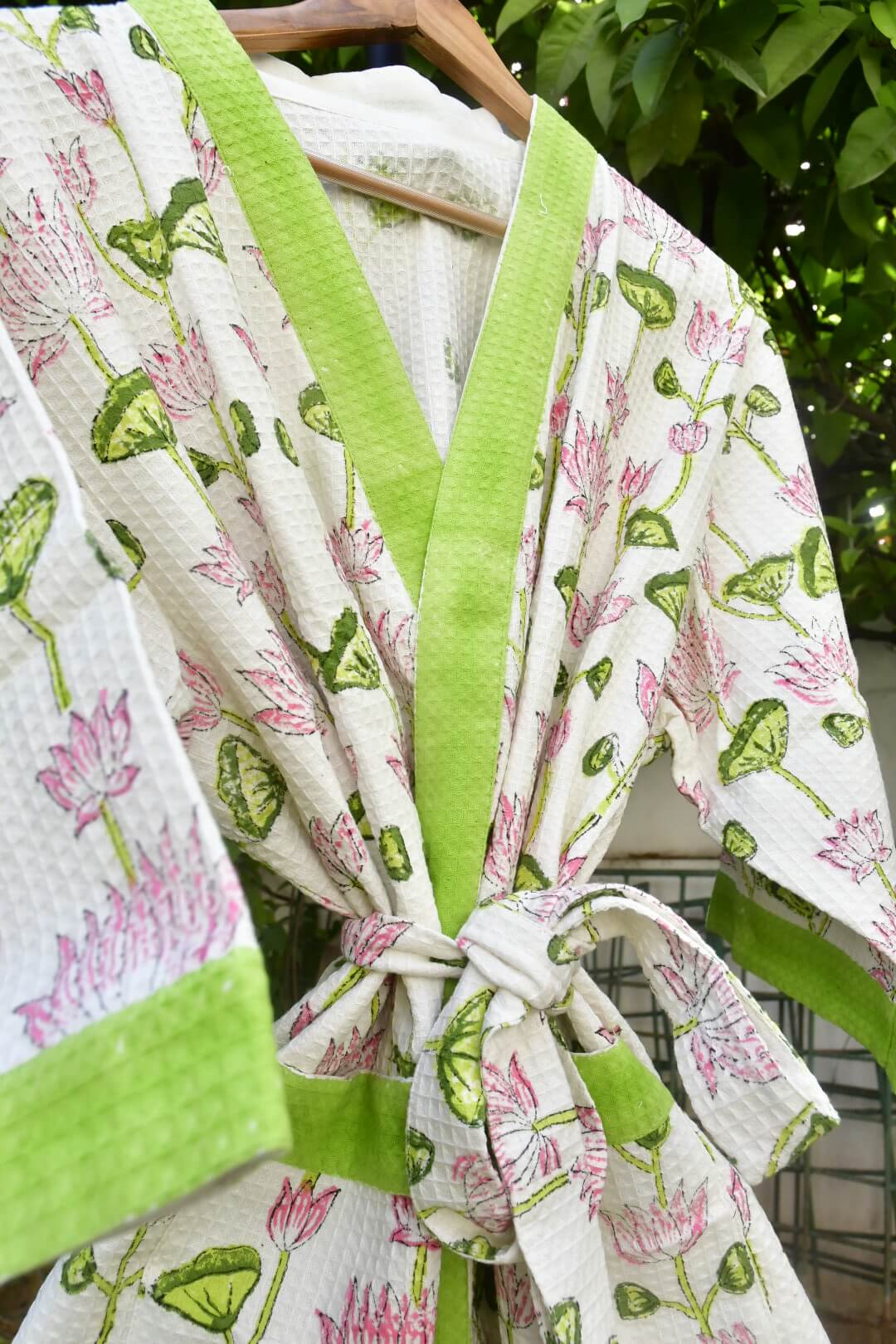 A white tie-front robe with a pink and green lotus print, featuring solid lime green trim and a matching tie belt, hanging on a wooden hanger against a background of green foliage.
