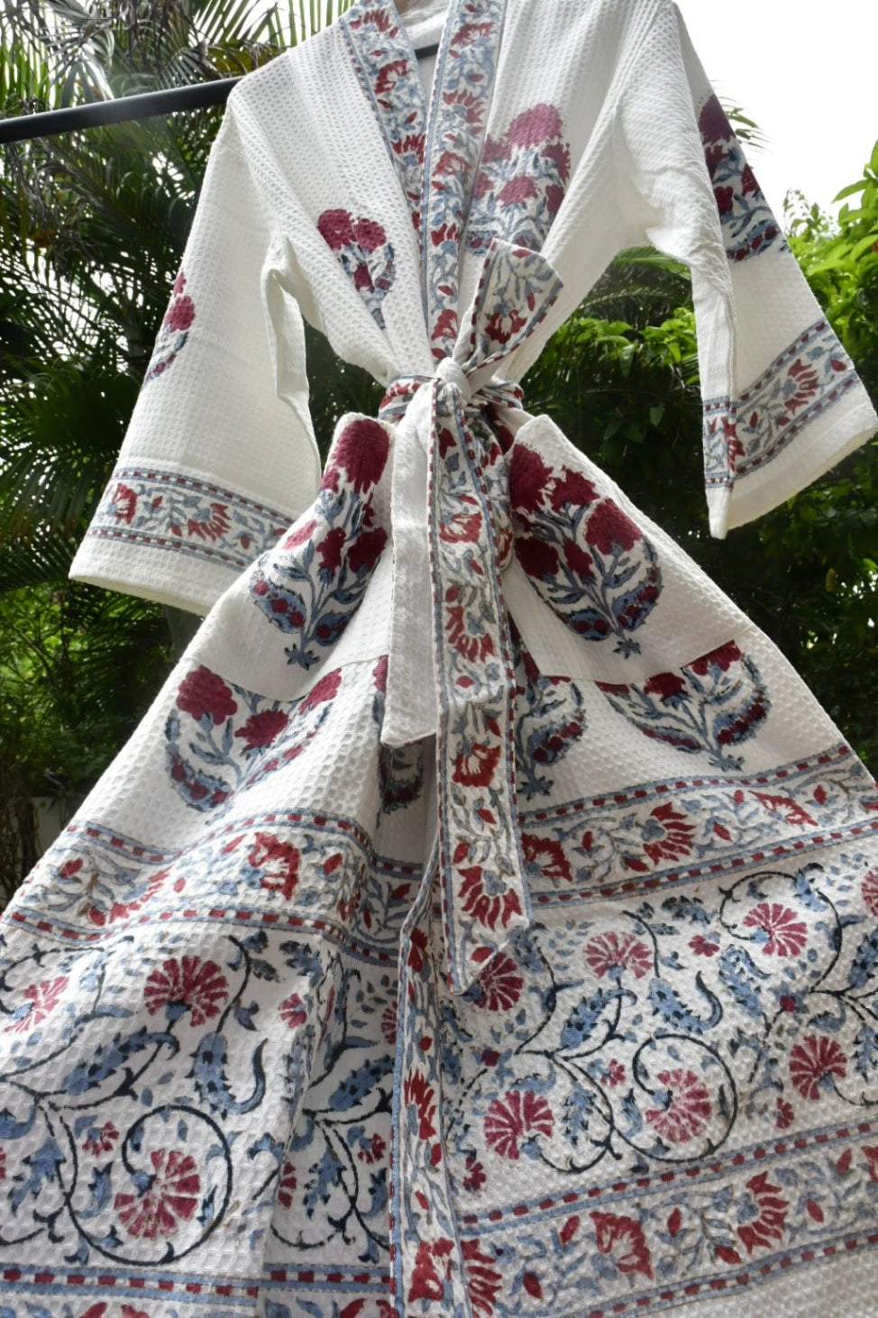 A white cotton block-print robe with red and blue floral motifs, hanging outdoors against a background of green foliage.