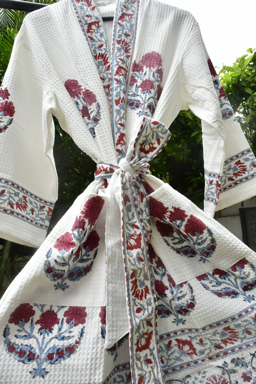 A white waffle-weave robe with red and blue floral block prints and a matching tie belt, shown hanging outdoors against a background of green foliage.