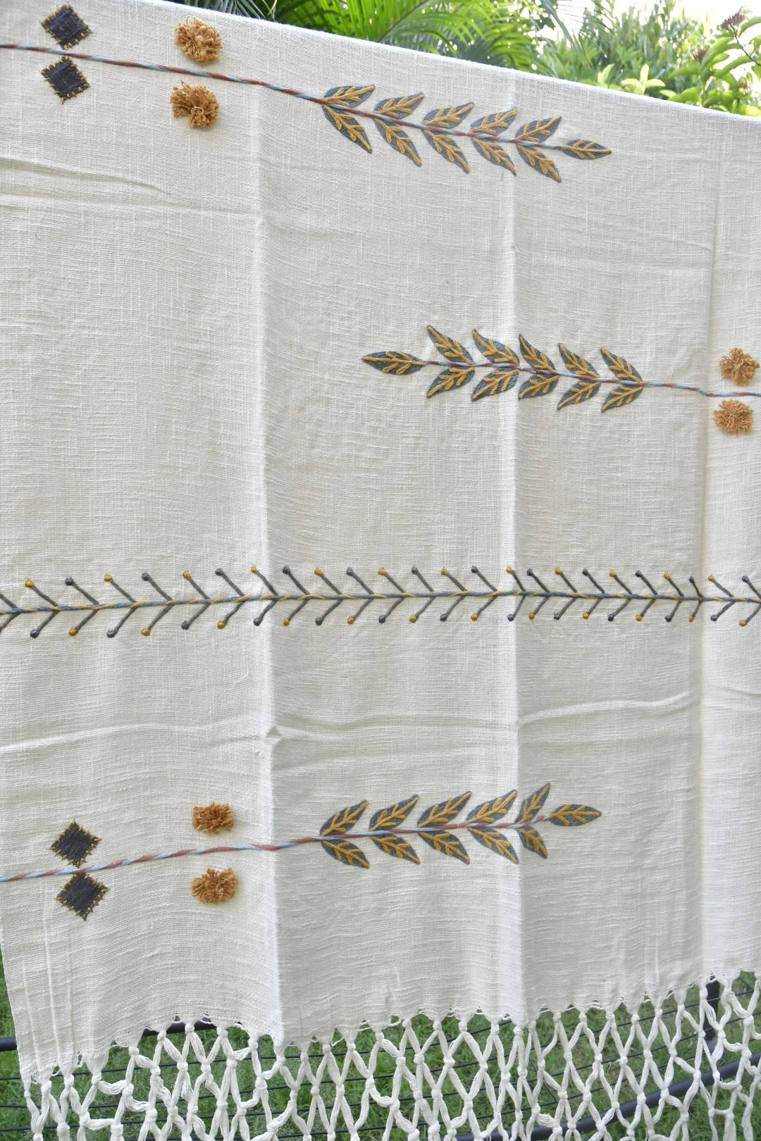An off-white linen textile, likely a tablecloth or decorative runner, featuring embroidered olive branches and small pom-poms in gold and brown, with a white woven fringe at the bottom, shown hanging outdoors.