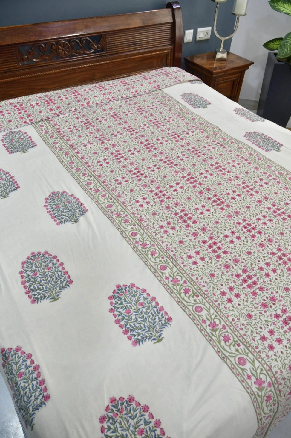 A cream-colored dohar / AC blanket with pink and green block-print floral motifs and borders, shown on a bed with a dark wood headboard in a bedroom setting.