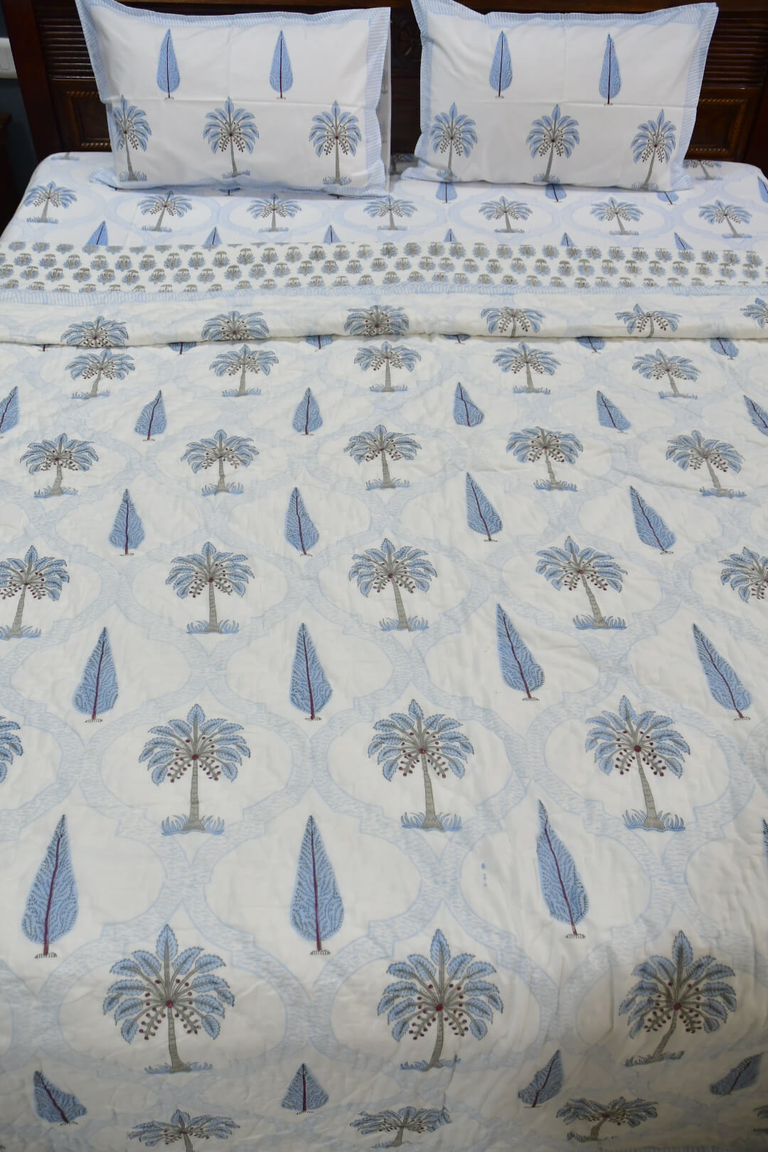 A bed made with a white quilt or duvet featuring a repeating blue palm tree and teardrop pattern, with matching pillowcases and a patterned border visible along the top.