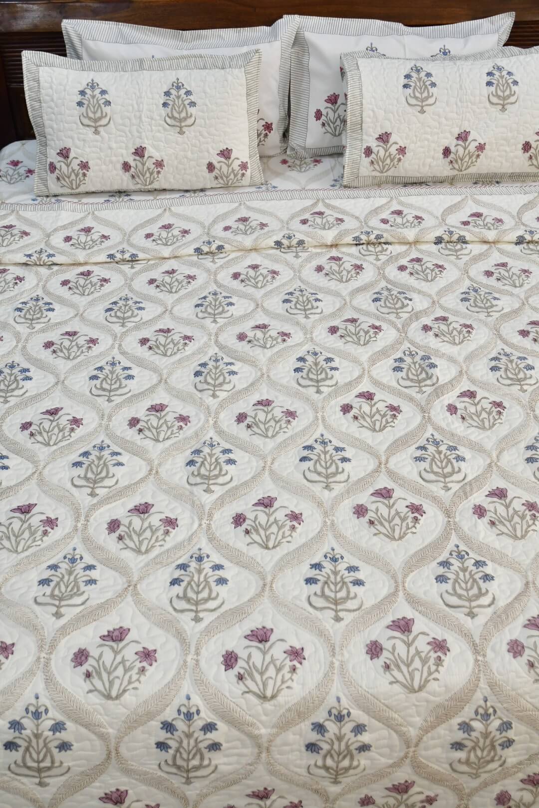 An off-white quilted bedspread with a repeating floral and geometric trellis pattern in beige, blue, and purple, styled on a bed with matching embroidered pillows.