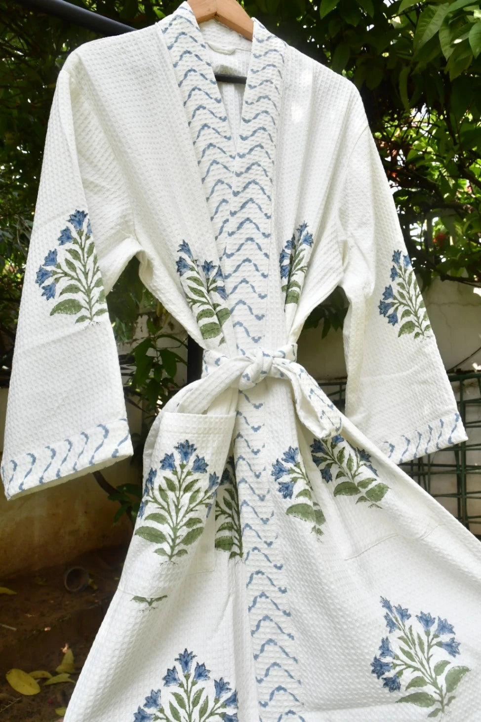 A white textured cotton robe with blue and green floral block prints and blue chevron patterns, hanging on a wooden hanger outdoors.