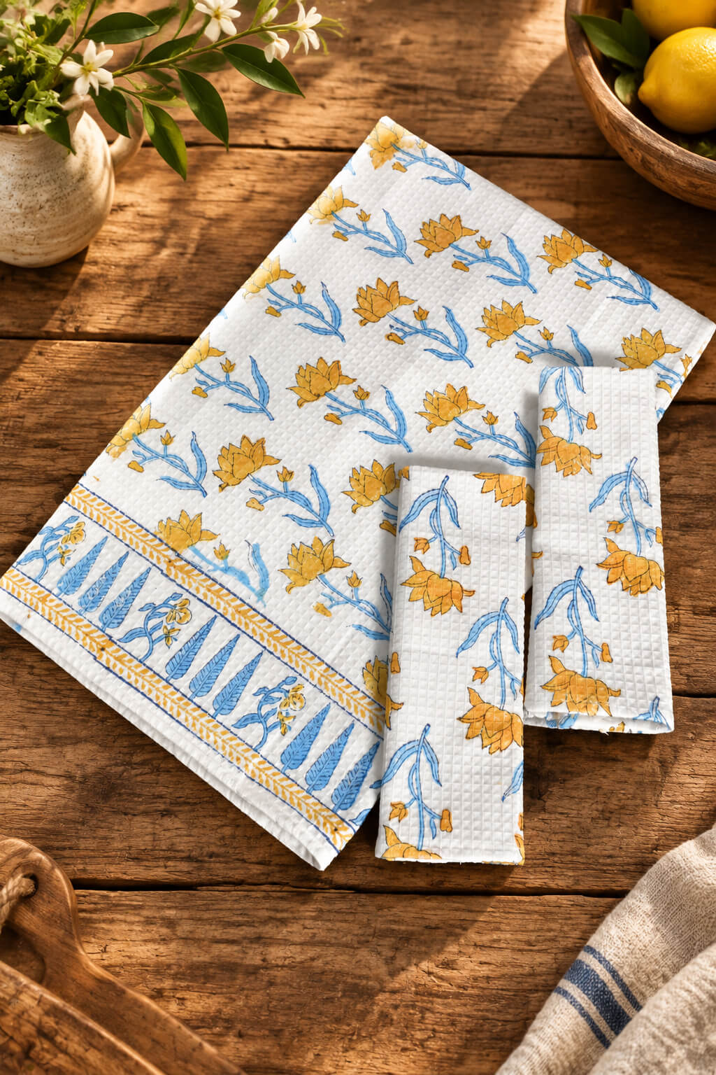 A set of three white cotton kitchen towels with a yellow and blue floral pattern and a striped border, arranged on a rustic wooden table with decorative props.
