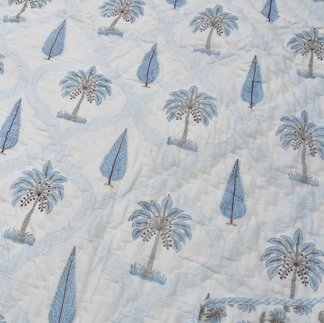 A close-up of a quilted rajai featuring a repeating pattern of blue palm trees and leaf motifs on a white background, with a folded corner showing a light blue and white striped border.