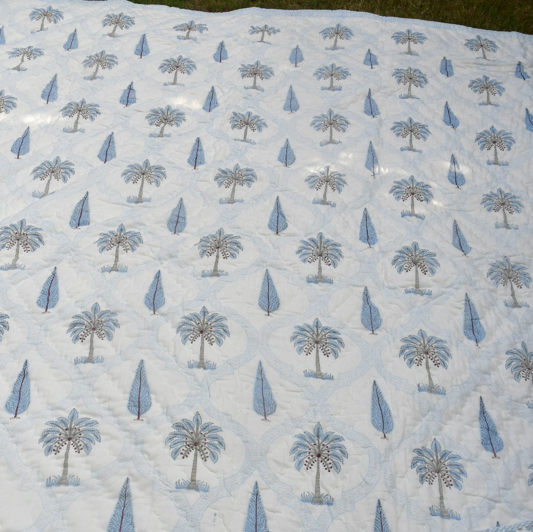 A close-up of a light blue and white printed quilt featuring a repeating palm tree and teardrop pattern, with a folded matching fabric piece placed in the upper right corner.
