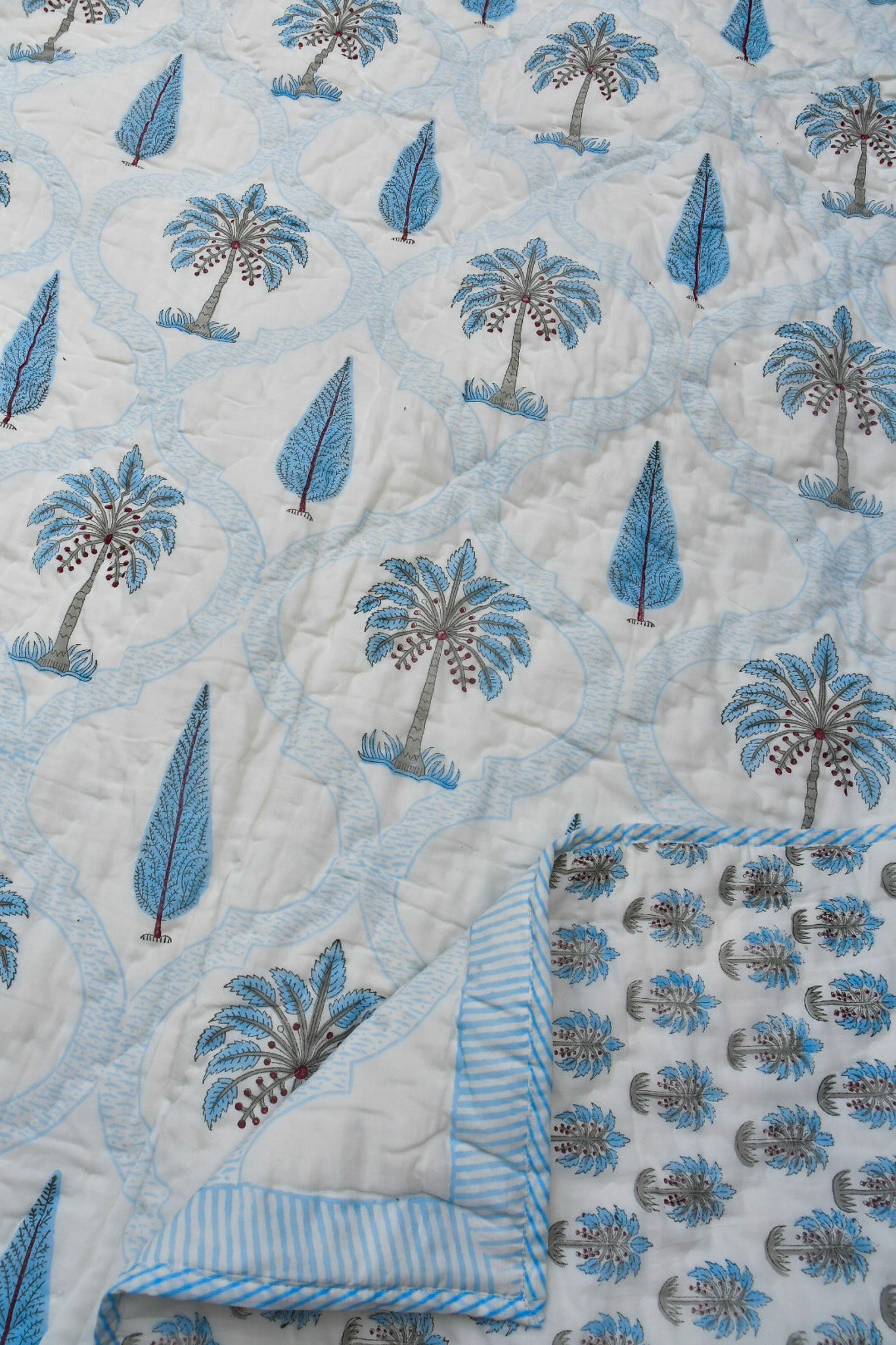 A close-up of a quilted blanket featuring a light blue and white tropical pattern with palm trees and leaf motifs, showing both the front design and the striped reverse side.