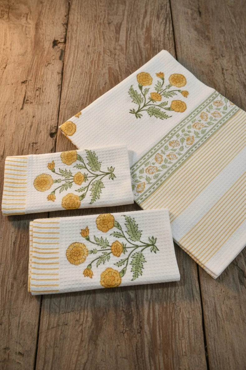 Three white waffle-weave kitchen towels with yellow floral and green leaf embroidery, arranged on a rustic wooden surface.