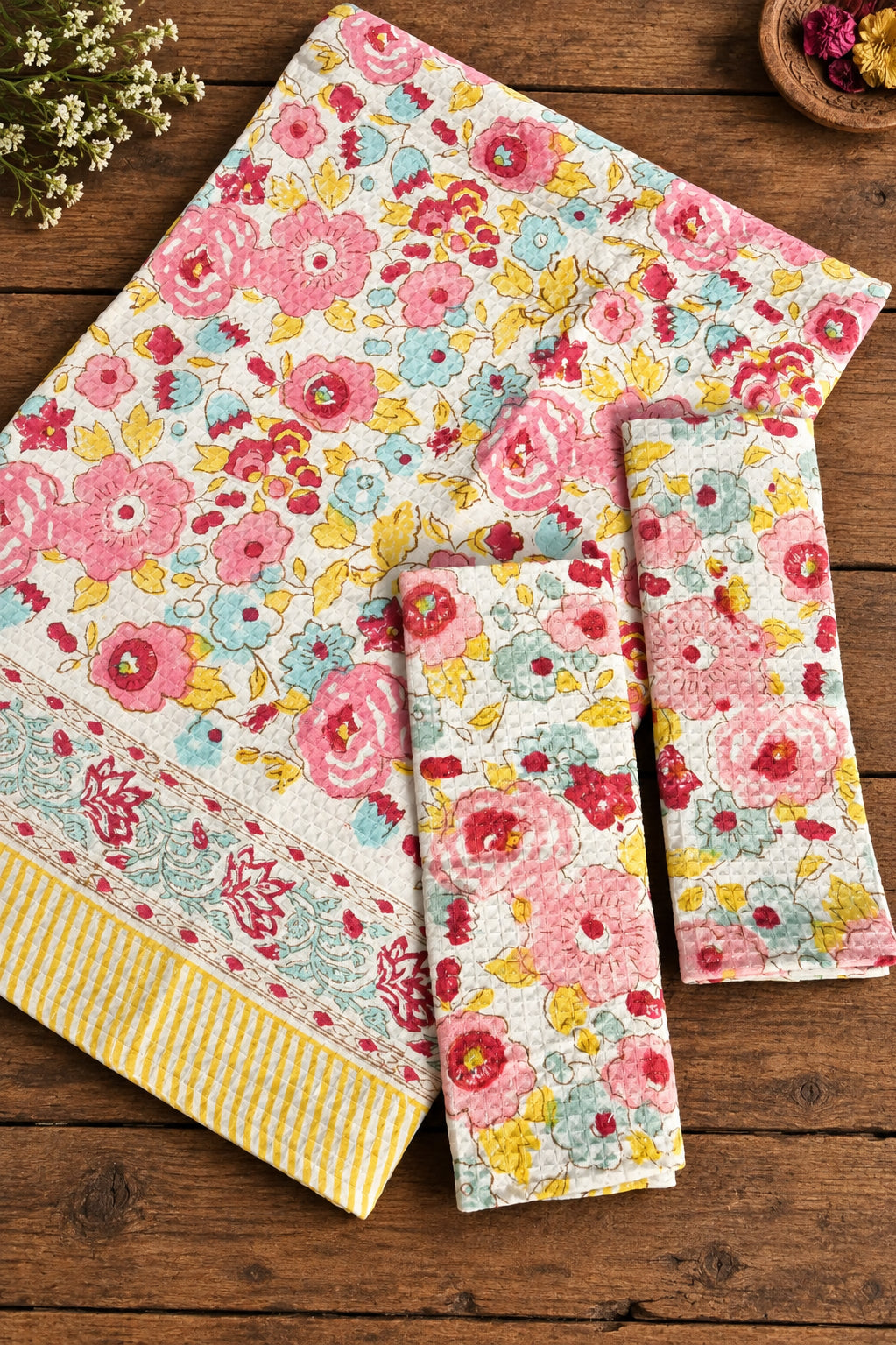 A floral fabric set laid out on a blue textured background, including a folded napkin or small tablecloth with pink, red, yellow, and blue flowers, plus two matching rectangular cloth pieces with striped and floral borders.
