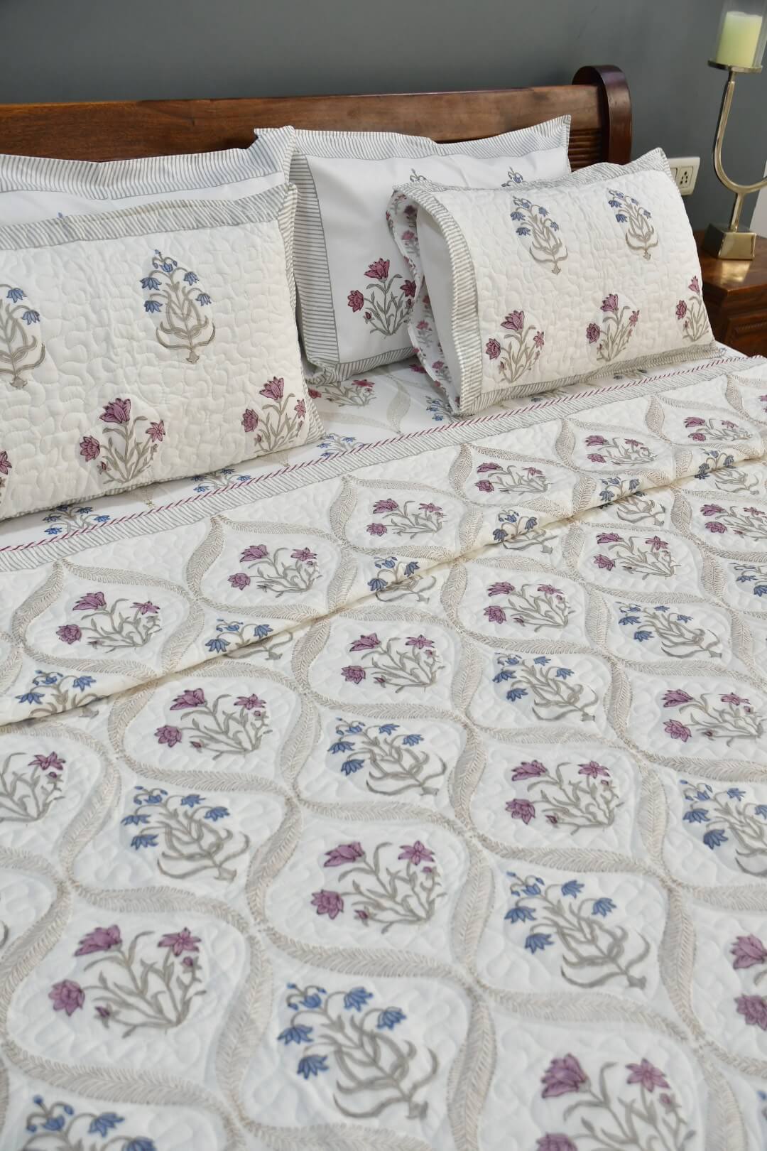 A quilted bedspread and matching pillow shams featuring a cream background with embroidered floral motifs in purple, blue, and green, styled on a bed with a wooden headboard.