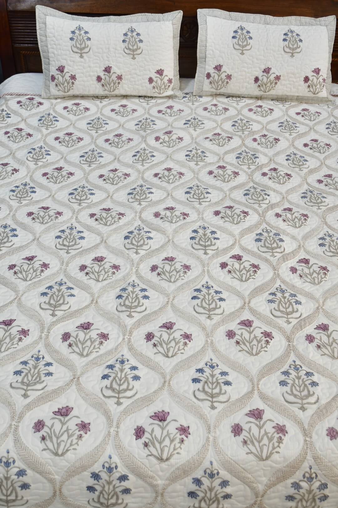 An quilted bedspread with matching pillowcases & bedsheet, featuring a cream background with a repeating geometric trellis pattern and floral motifs in purple, blue, and beige.