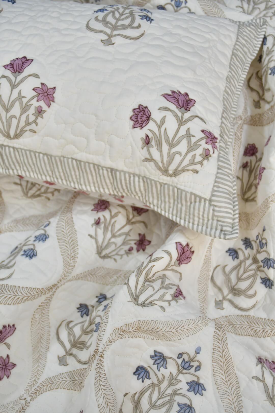 A close-up of a quilted bedding set featuring a cream-colored quilt and matching pillowcase with embroidered floral motifs in dusty pink, blue, and beige.