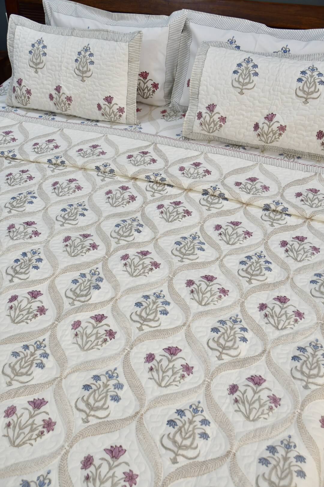An off-white quilted bedspread with a repeating floral block-print pattern in purple, blue, and beige, styled with matching pillowcases on a bed.