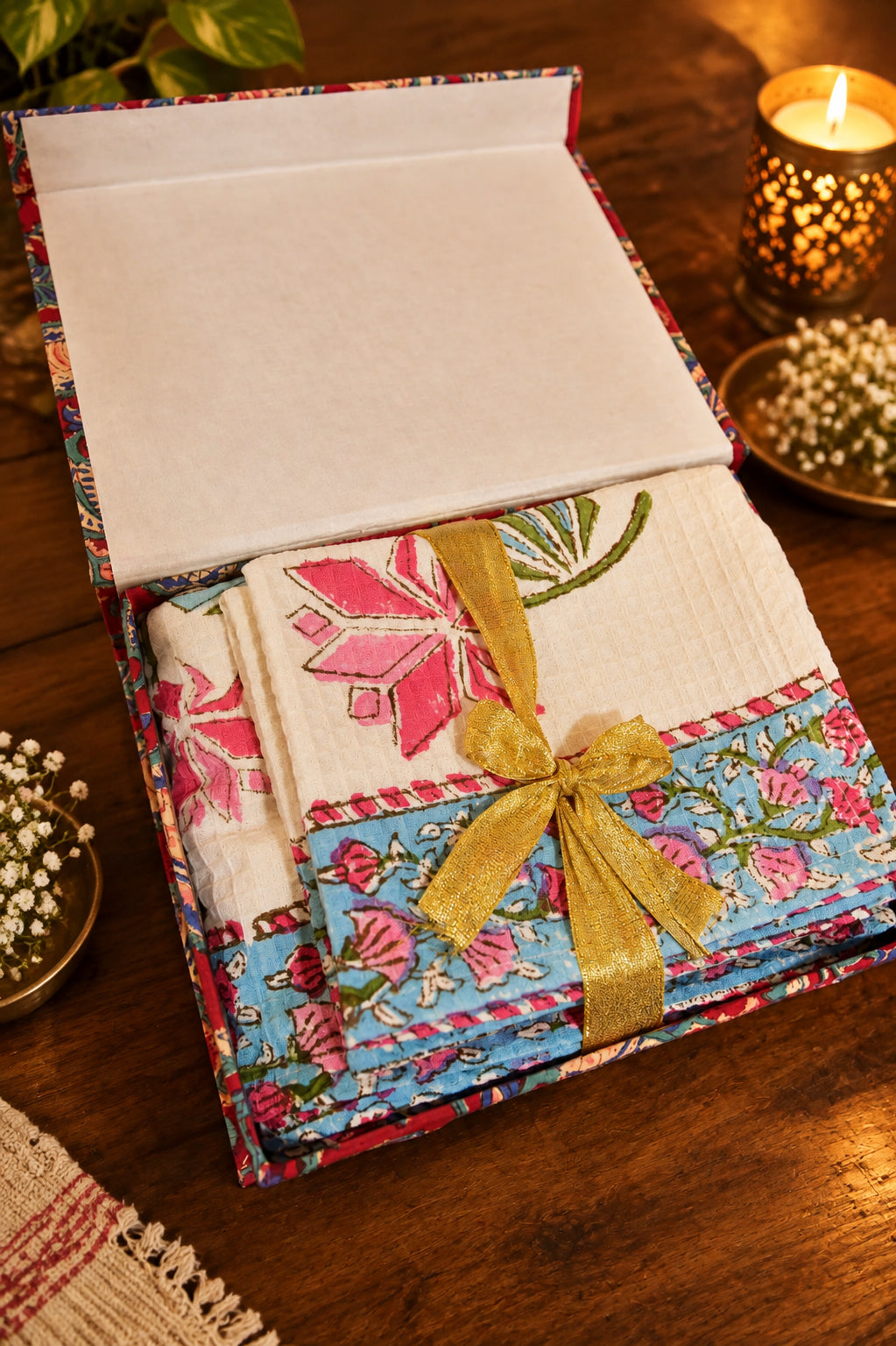 A folded textile gift set in a patterned box with a white interior, tied with a gold ribbon. The fabric features a white textured section with pink floral embroidery and a blue floral border.