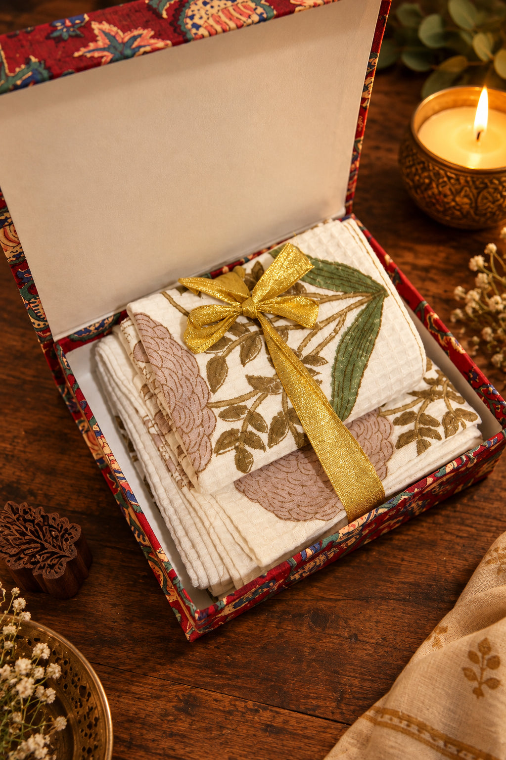 A gift box of folded embroidered cotton textiles, likely dupattas or scarves, tied with a gold ribbon. The textiles are white with brown and green floral embroidery, and the box has a red patterned border.