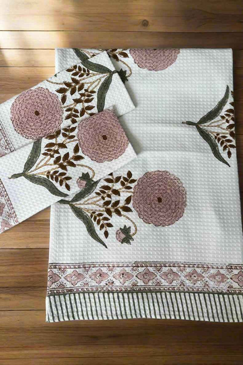 A white textured cotton tablecloth with a floral block print pattern in dusty pink and brown, laid flat on a wooden floor alongside matching napkins.