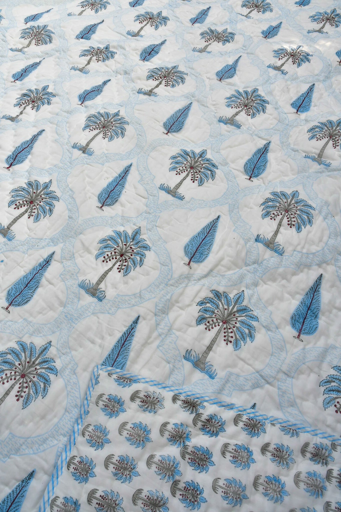 A close-up of a quilted jaipuri design featuring a repeating pattern of blue palm trees and blue teardrop motifs on a white background, with a matching blue and white border visible in the bottom corner.