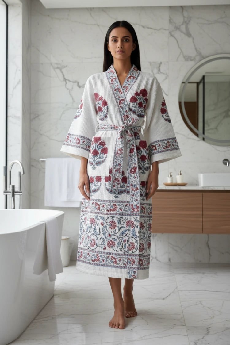 A woman stands barefoot in a modern bathroom wearing a white cotton robe with red and blue floral patterns and a matching tie belt.