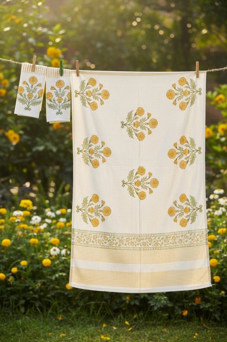 A cream-colored cotton towel with yellow and green floral block prints hangs on a clothesline outdoors in a garden with yellow flowers.