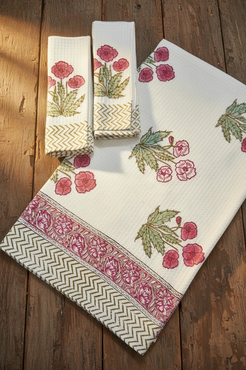 A set of white cotton kitchen towels with pink floral prints and green leaves, featuring decorative borders, displayed on a rustic wooden background.