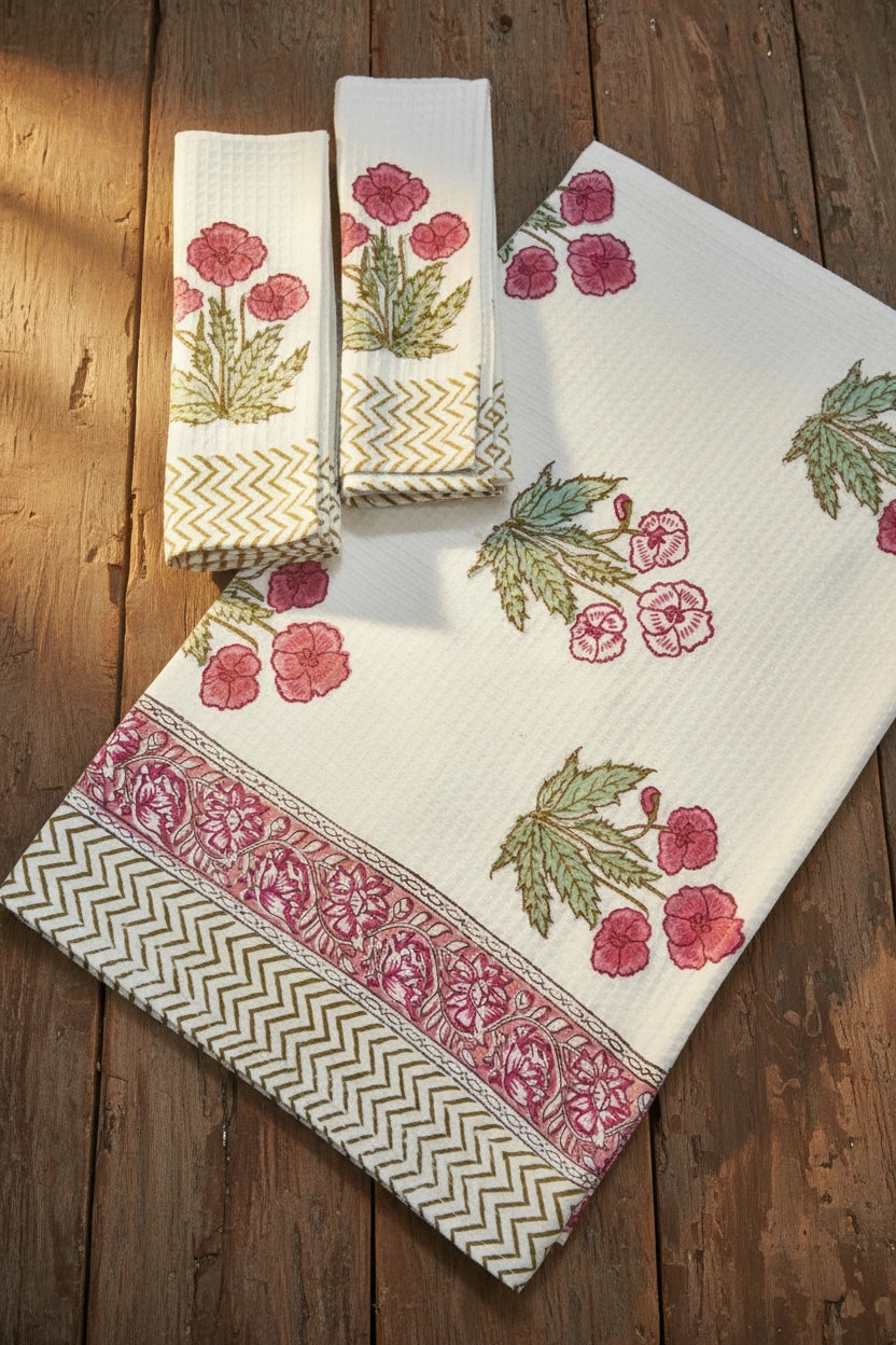 A set of white cotton kitchen towels with pink floral prints and green leaves, featuring decorative borders, displayed on a rustic wooden background.