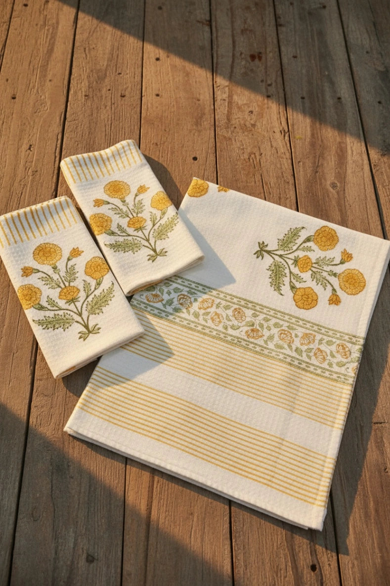 Three cream-colored cotton kitchen towels with yellow and green floral embroidery and striped borders, arranged on a rustic wooden deck with warm sunlight and shadows.
