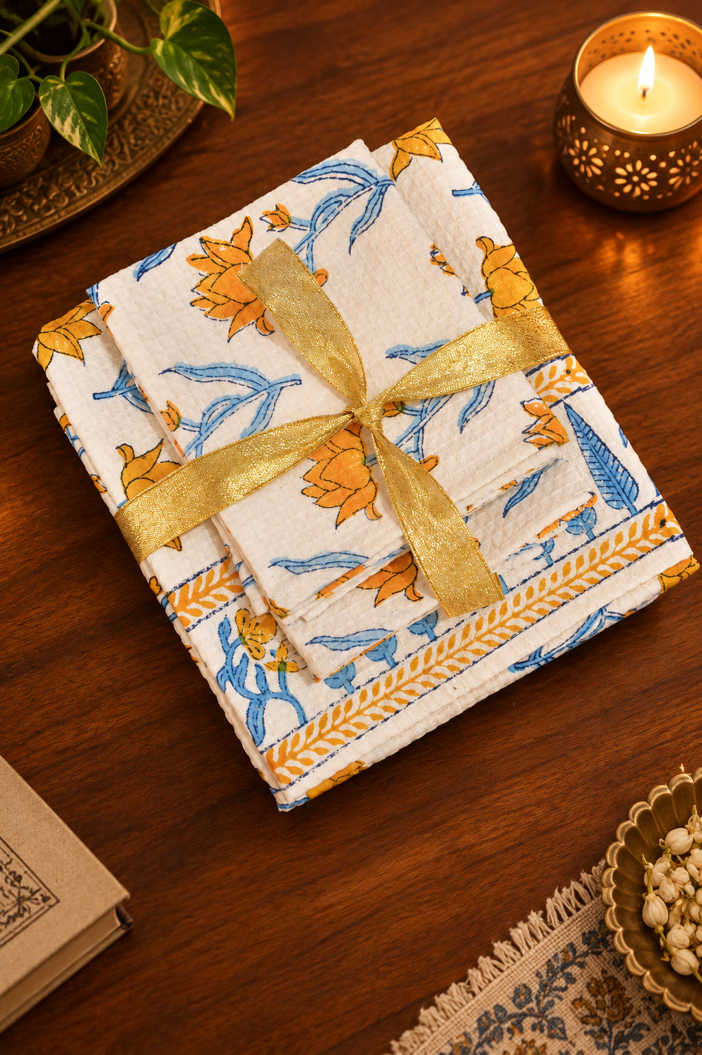 A stack of folded white cotton napkins or cloths with a blue and orange floral print, tied with a shiny gold ribbon, displayed on a dark wooden table with decorative props including a lit candle, a book, and a small bowl.