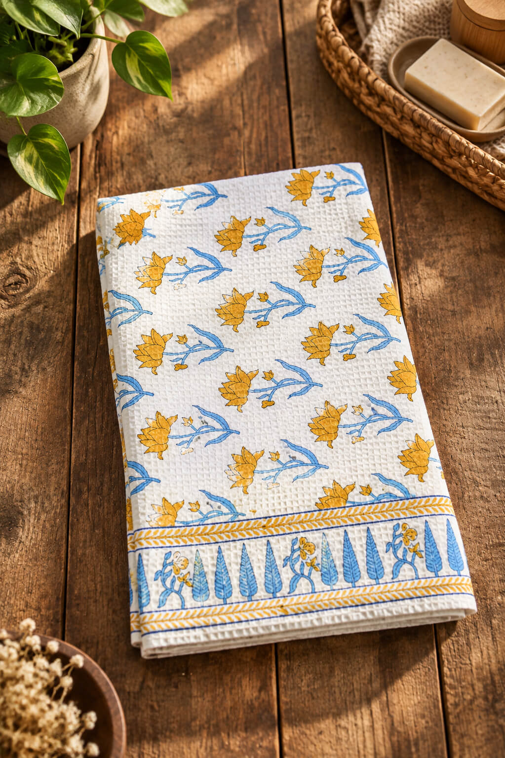 A folded white cotton kitchen towel with a blue and yellow floral and bird pattern, displayed on a rustic wooden table with decorative props.