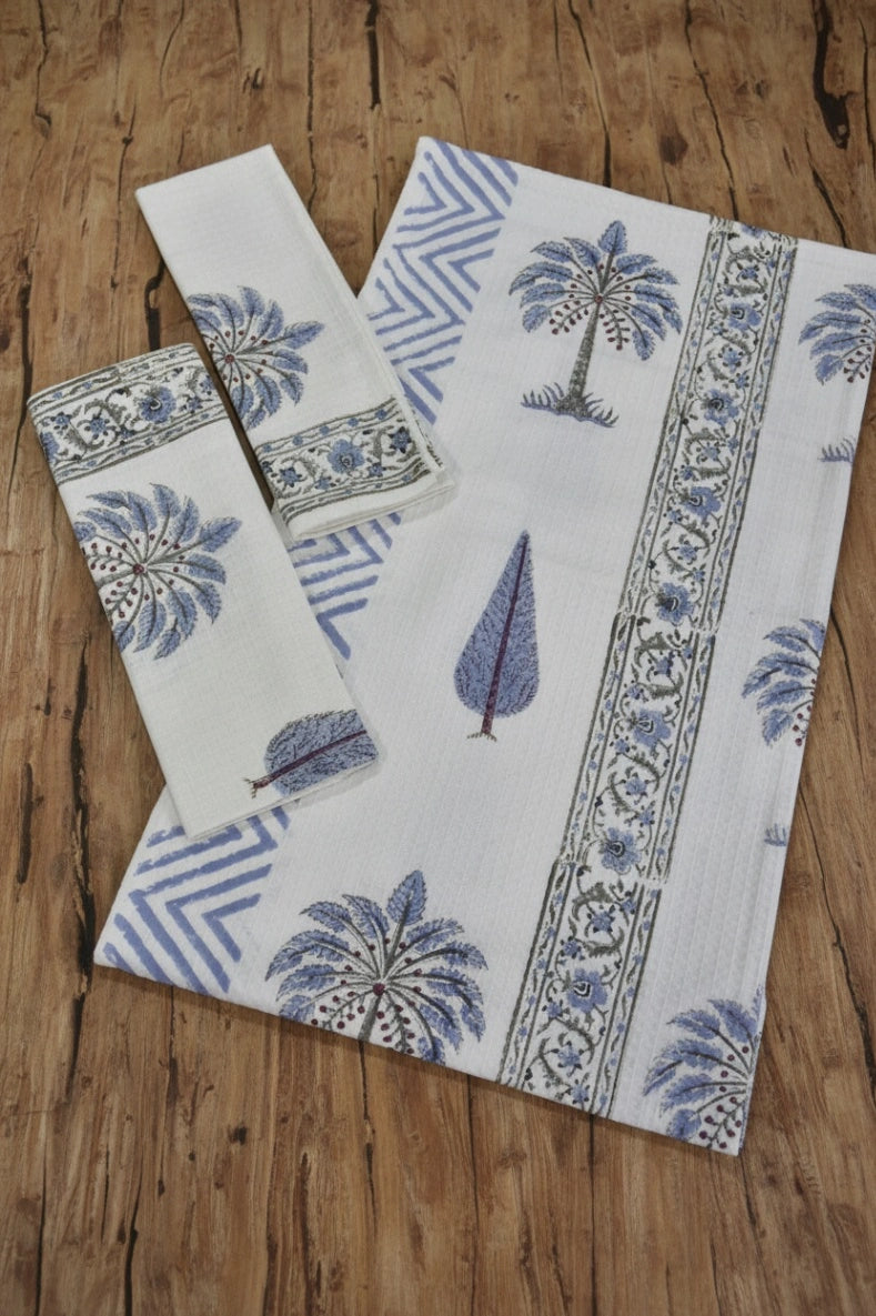 A set of white cotton towels, including a large rectangular cloth and two smaller square pieces, printed with blue palm tree motifs, floral borders, and geometric patterns, arranged on a rustic wooden surface.