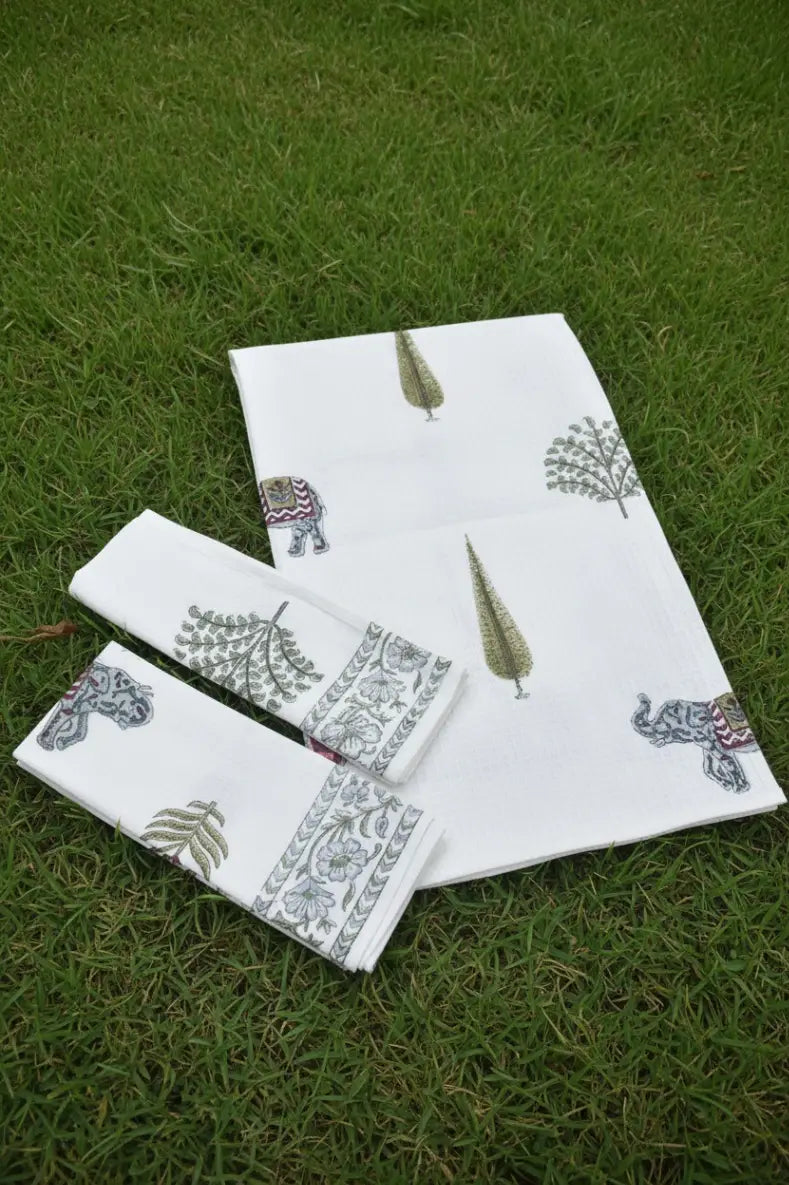 A set of white cotton towels with hand-painted elephant and tree motifs, arranged on a green grass lawn.