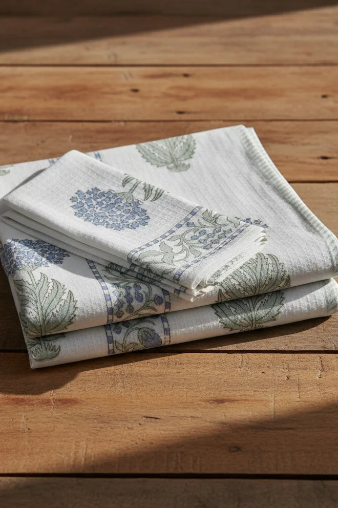 A stack of folded cotton towels with a blue and green botanical block print pattern, displayed on a rustic wooden surface.