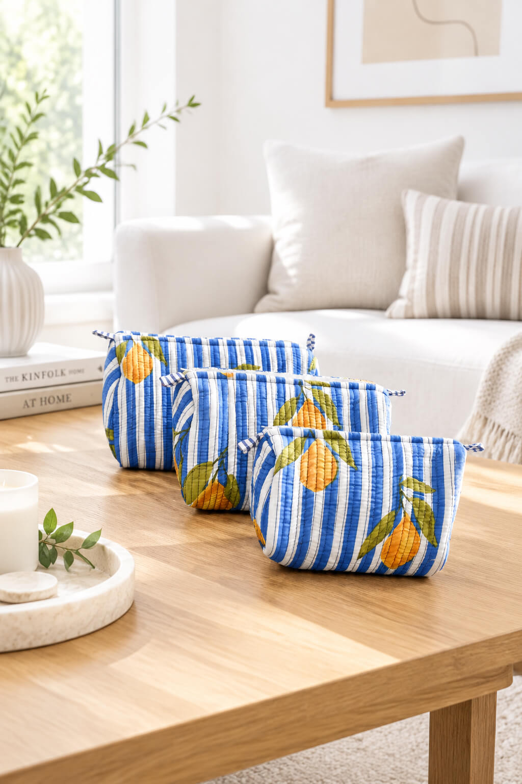 Three zippered pouches with blue and white stripes and a lemon print, arranged on a light wood table in a bright, modern living room setting.
