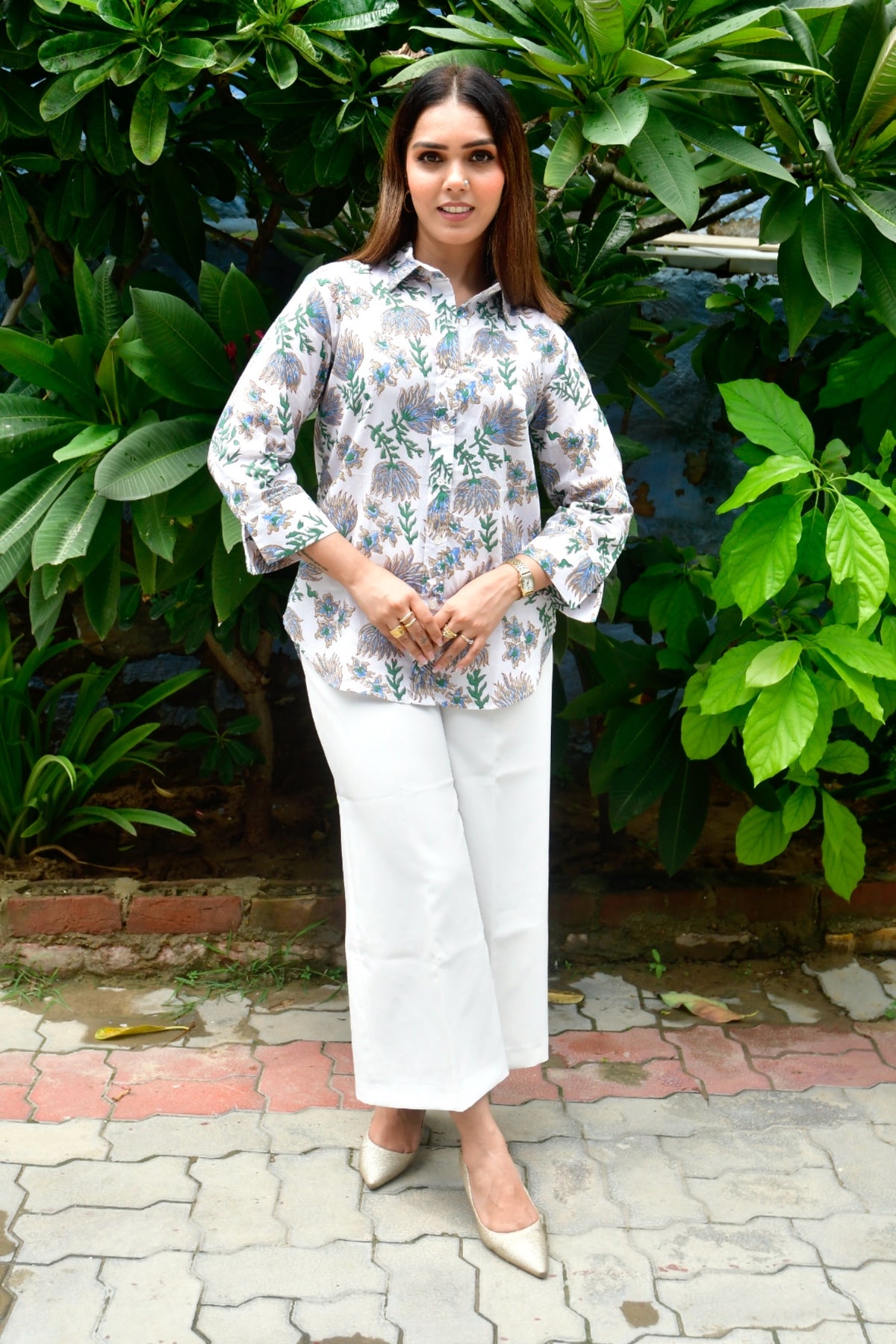 Woman in a floral blouse and white pants standing in front of green foliage.