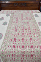 A floral patterned dohar / AC blanket with pink and green motifs on a cream background, laid out on a bed with a dark wooden headboard.