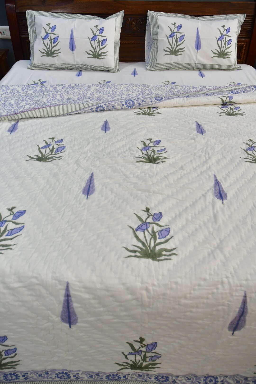 A white quilted bedspread set on a bed, featuring embroidered blue and green floral motifs and scattered blue teardrop shapes, with two matching pillowcases at the head.