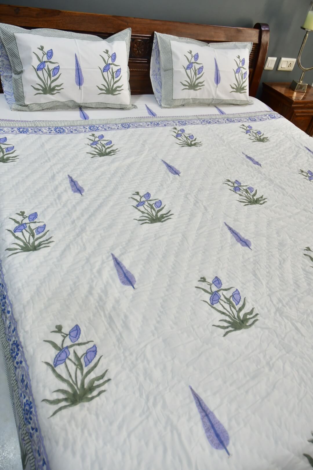 A white quilted bedspread with blue and green floral motifs and matching pillowcases, styled on a bed with a wooden headboard in a bedroom setting.